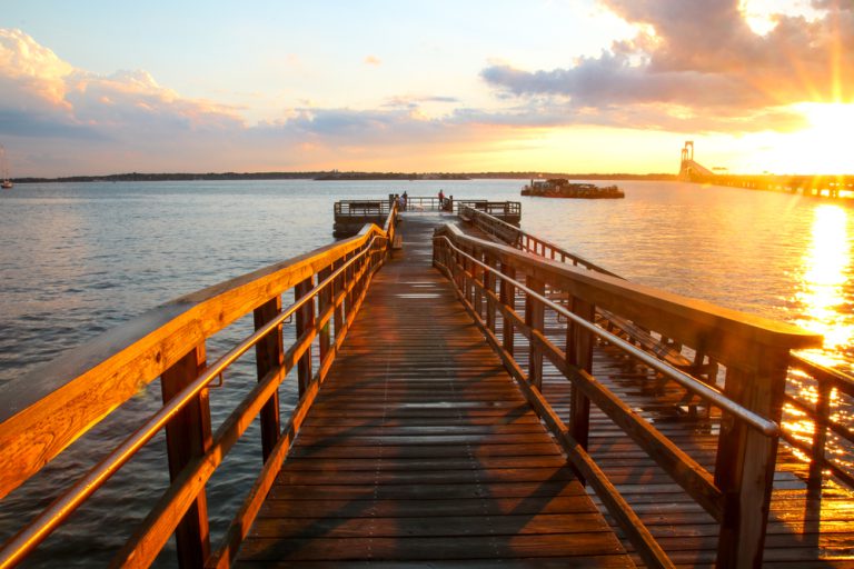The sun rises and turns the water by a wooden pier golden. Several people stand, fishing, at the edge of the pier.