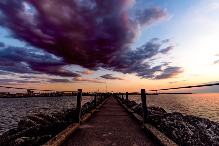 A dramatic sunset turns the sky purple and orange over a harbor, a long paved pier with rocky edges leading out into the water.