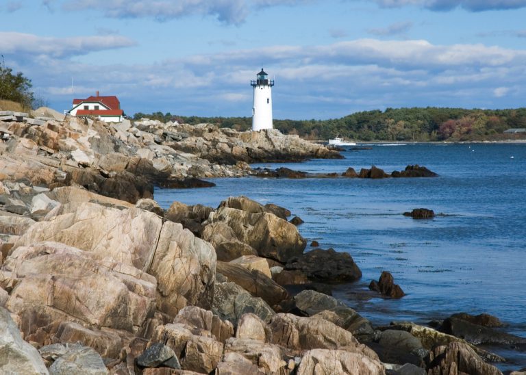 A white lighthouse stands on the rocky shore next to deep blue water, a tug boat sailing in the distance. The shore is surrounded with densely forested banks under a blue sky with white clouds.
