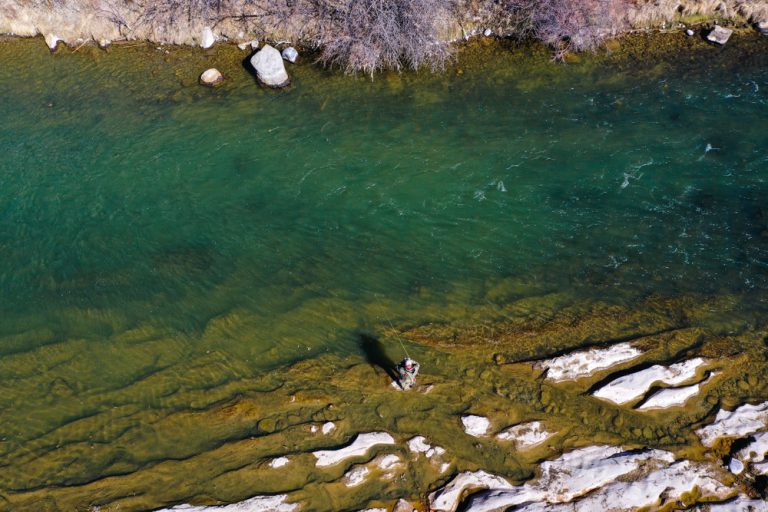 An angler wading and fishing in a calm, blue-green river surrounded by red scrub brush and a brown, rocky mesa.