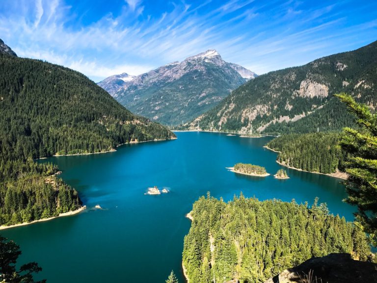 A bright blue lake sits between steep mountains covered with evergreen trees and rocky ridges.
