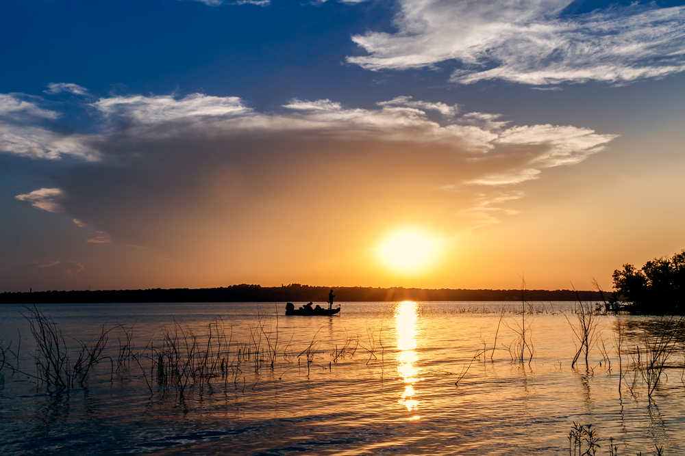 A man stands in the prow of a boat casting a fishing line. The boat is in the middle of a deep lake, and the sun is setting over the horizon.