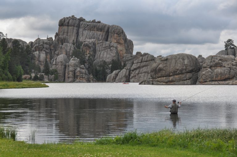 A man stands in the middle of a lake, casting a fishing pole toward deeper waters. Rocky cliffs make up the other bank.