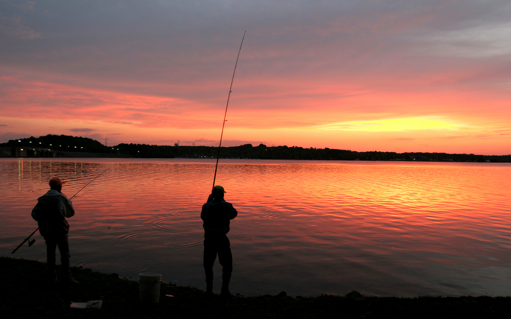 Two people with long fishing poles silhouetted on the shore of a lake, as the sun rises behind distant clouds.