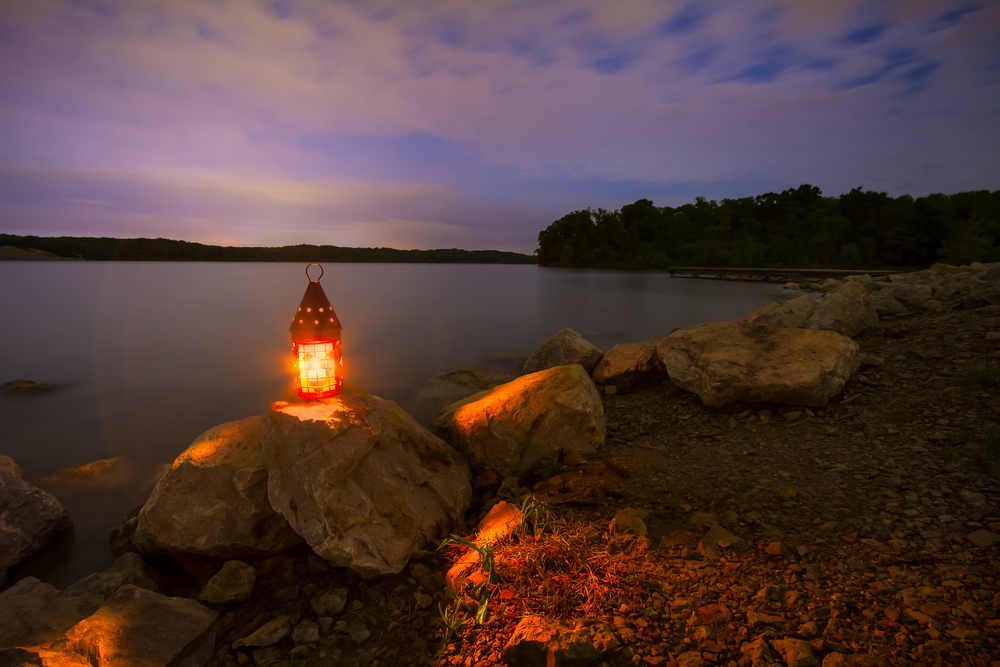 A lantern atop a large rock on a lake shoreline glows bright orange under a purple sky.