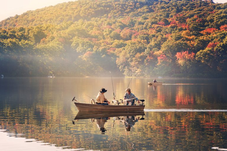 Two boats, one with two people and another with one person, sit on a calm lake, fishing. The bank is surrounded by dense forest.