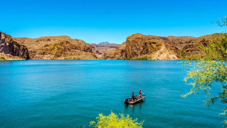 Two men stand, fishing, on a boat in the middle of a bright blue lake surrounded by rocky hills under a blue sky.
