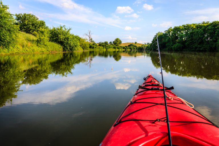 A fishing pole leans out of a red kayak on a calm lake surrounded by dense green trees.