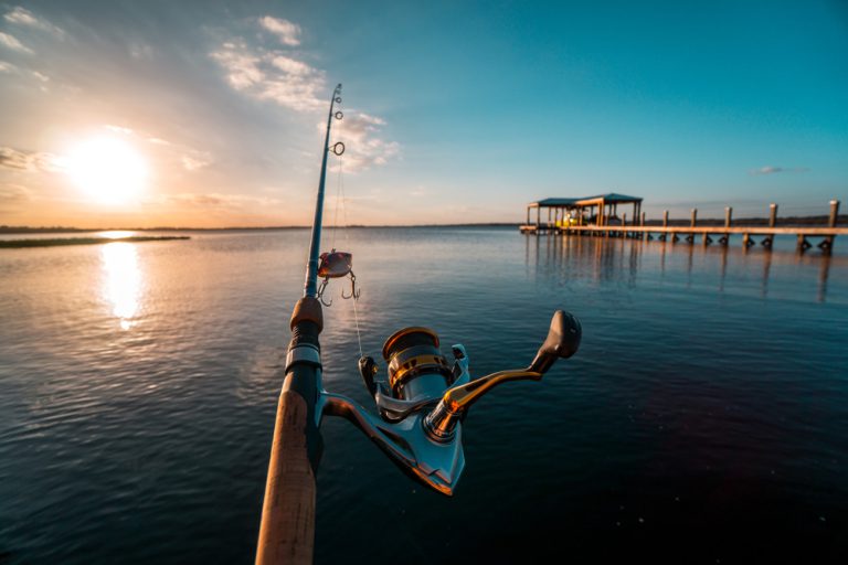 A cast fishing pole in a calm body of water, next to a long wooden dock. The sun sets over the horizon