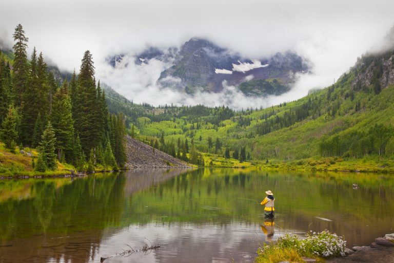 A person in a yellow jacket standing in a lake tosses a fly fishing line. The lake is surrounded by green trees, and clouds surround the distant mountains.
