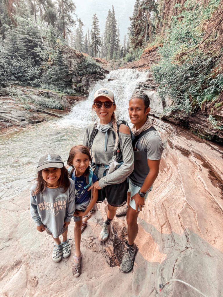 Family of four - mom, dad, son, and daughter - stand in front of waterfall at Glacier National Park