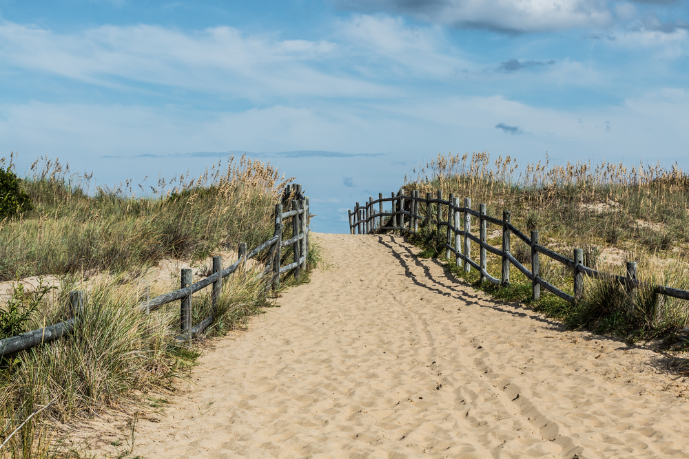 A sandy beach pathway bordered by wooden railings and grass covered dunes leads to the ocean seen in the distance.