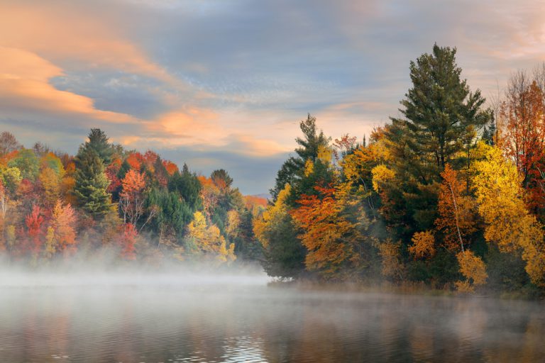 Fog hovers over a lake surrounded by orange, yellow and green trees under a pale orange and blue sky.