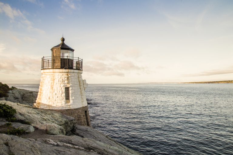 A white painted brick lighthouse on the edge of a gray rocky cliff overlooking the sea.