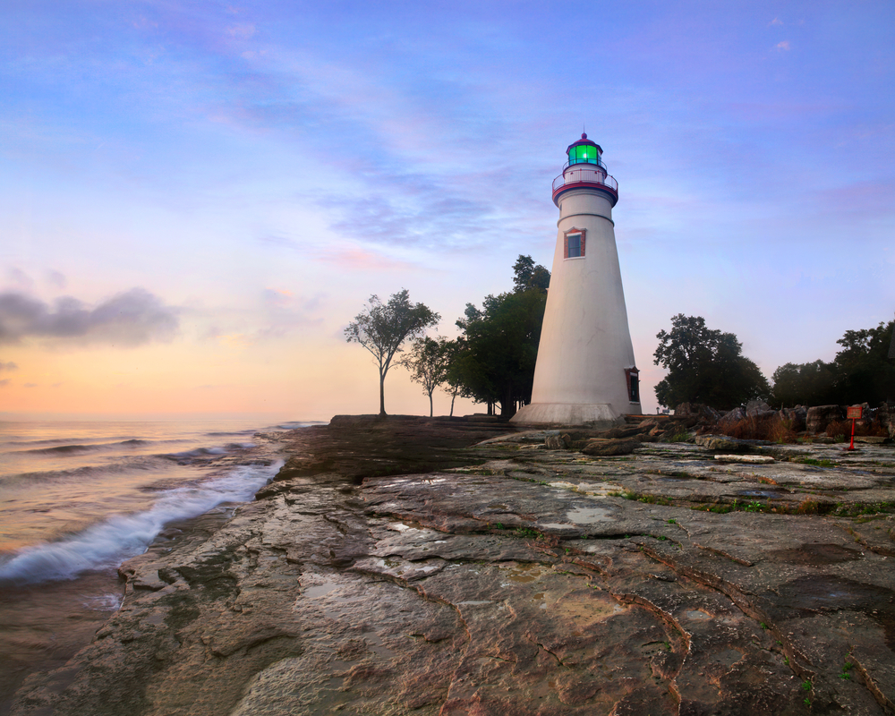 A white and red lighthouse on top of a rocky cliff shoreline. Trees grow beside the lighthouse under a pale pink and blue sky.