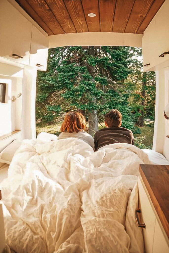Couple lays in bed looking out the back of their open campervan doors