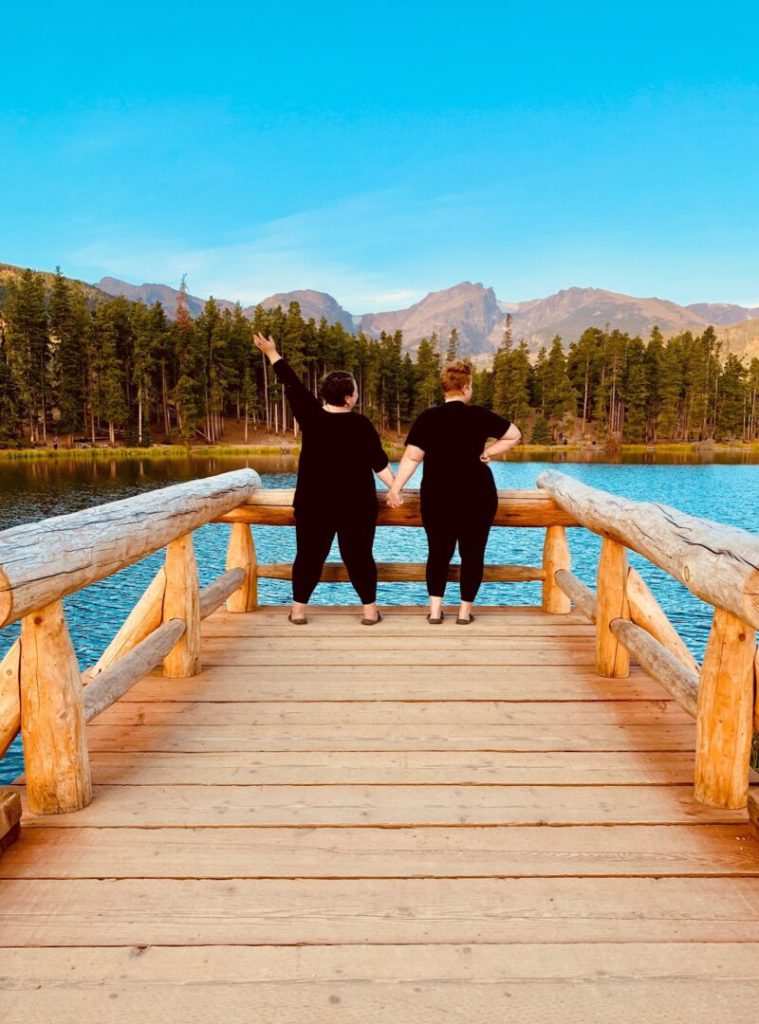 Women stand at the end of a pier holding hands looking over a lake