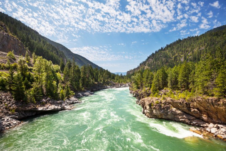 A blue-green river rushes through a valley of rocky mountains covered in tall green trees.