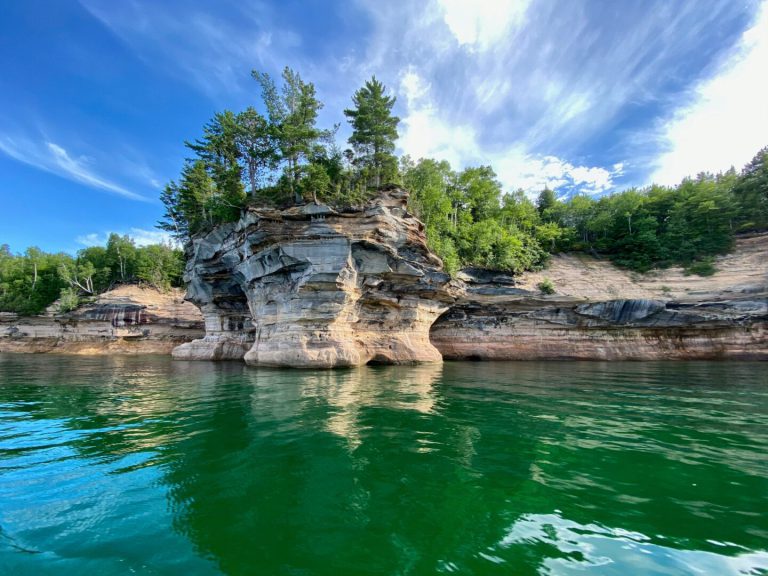 Green and blue waters roll beneath a large rock formation with green trees growing on top