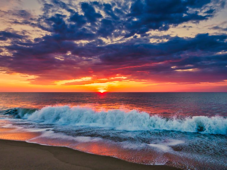 The sun rises over the ocean. Waves crash into the sandy shore.