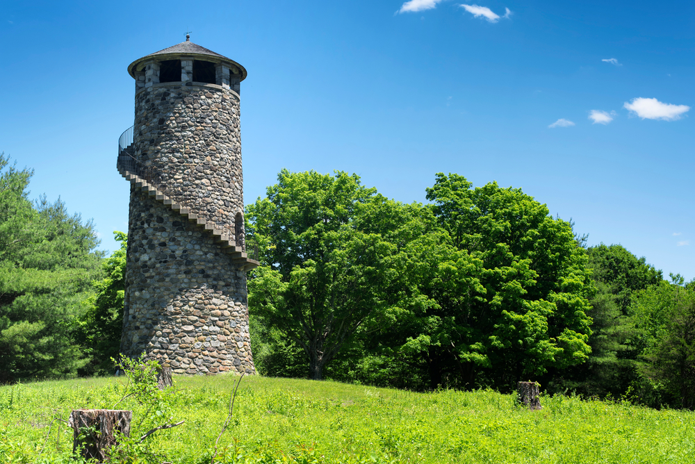 A stone tower with narrow stairs wrapped around it sits in a grassy meadow next to green trees with a blue sky overhead.