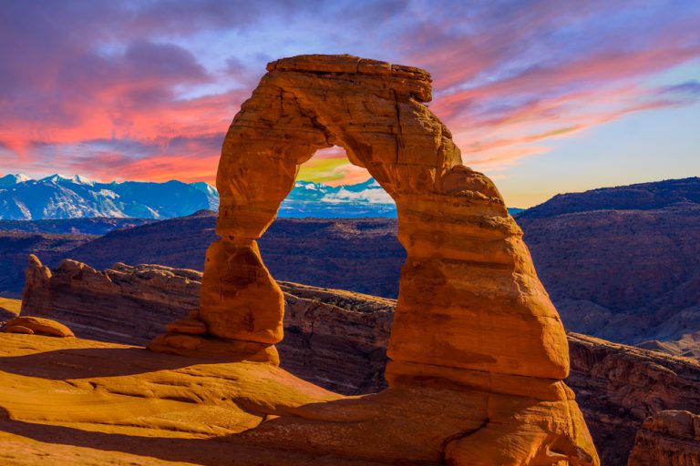 A red-rock arch sits in front of red-rock cliffs and snowy mountains in the distance with pink and orange clouds overhead