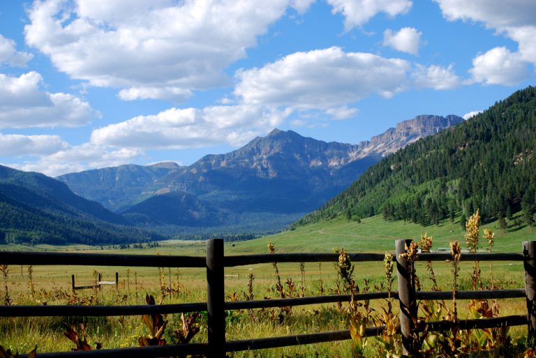 A wooden fence in a grassy field. Tall mountains stand in the background under puffy white clouds in a blue sky