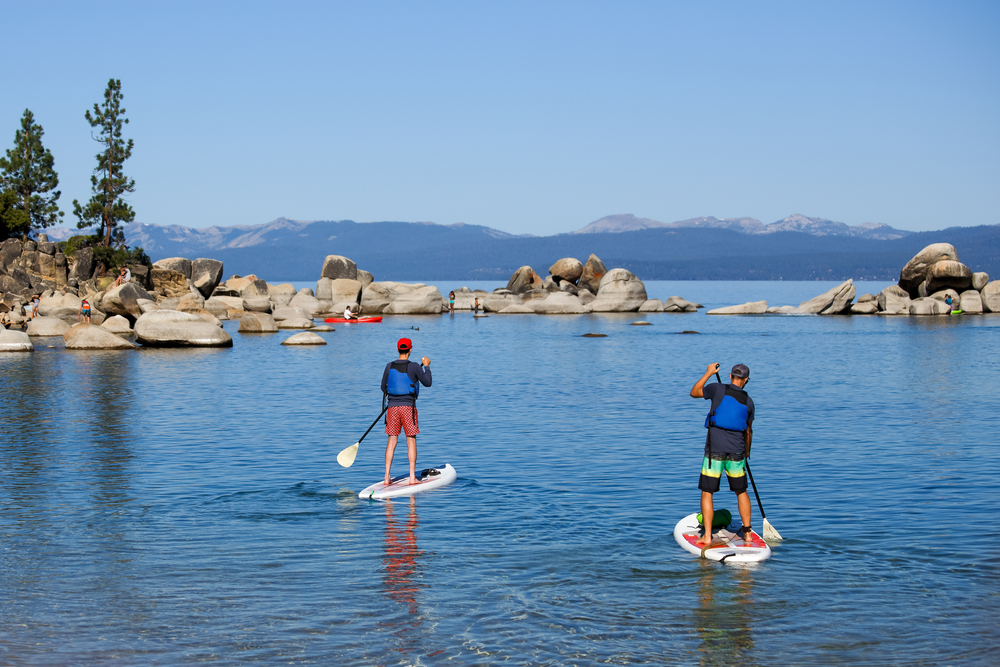 Two young men wearing life jackets paddle boarding on a lake with a mountain and boulder backdrop.