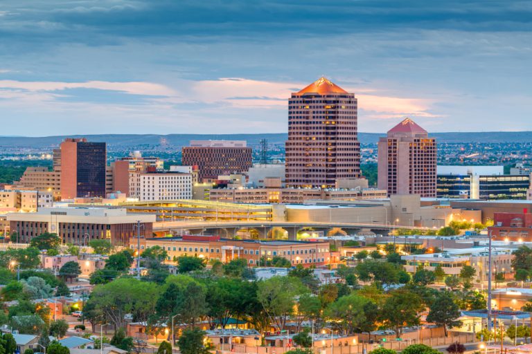 A city lit up at dusk. Green trees surround buildings in the foreground. A small mountain range can be seen in the distance.