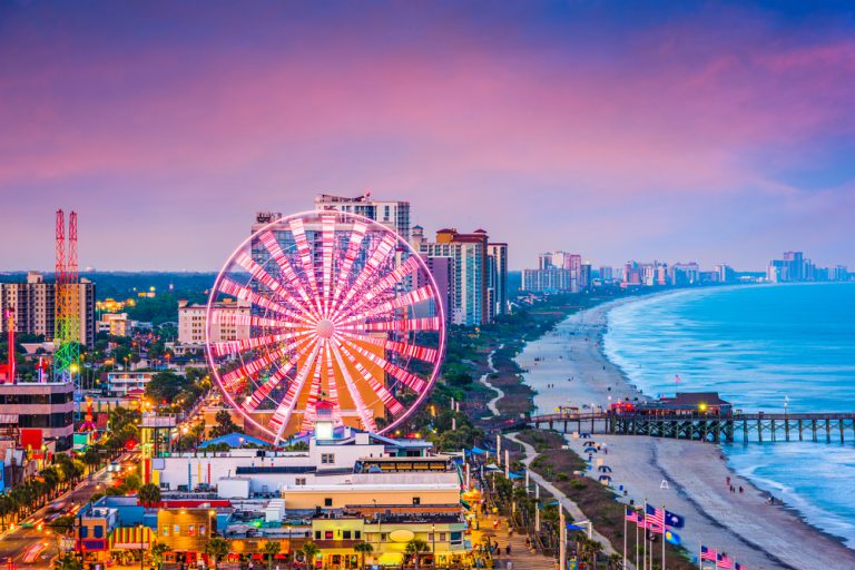 A Ferris wheel and numerous tall buildings line a sandy beach under a purple sky at sunset.