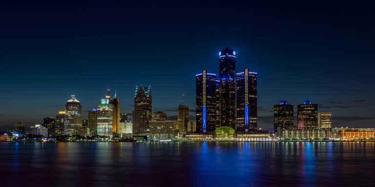 Bright lights shine in the buildings of a metropolitan skyline at night, reflected in a calm, black body of water.