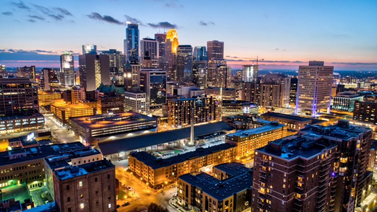 A downtown skyline glows with lights illuminating its tall buildings at dusk.
