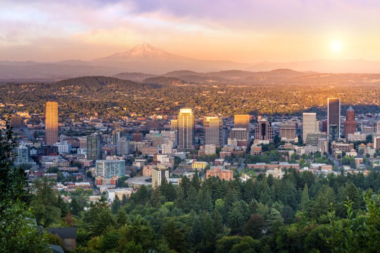 The downtown Portland skyline at dusk is dominated by the looming Mt. Hood in the background under a pale purple and golden sky.