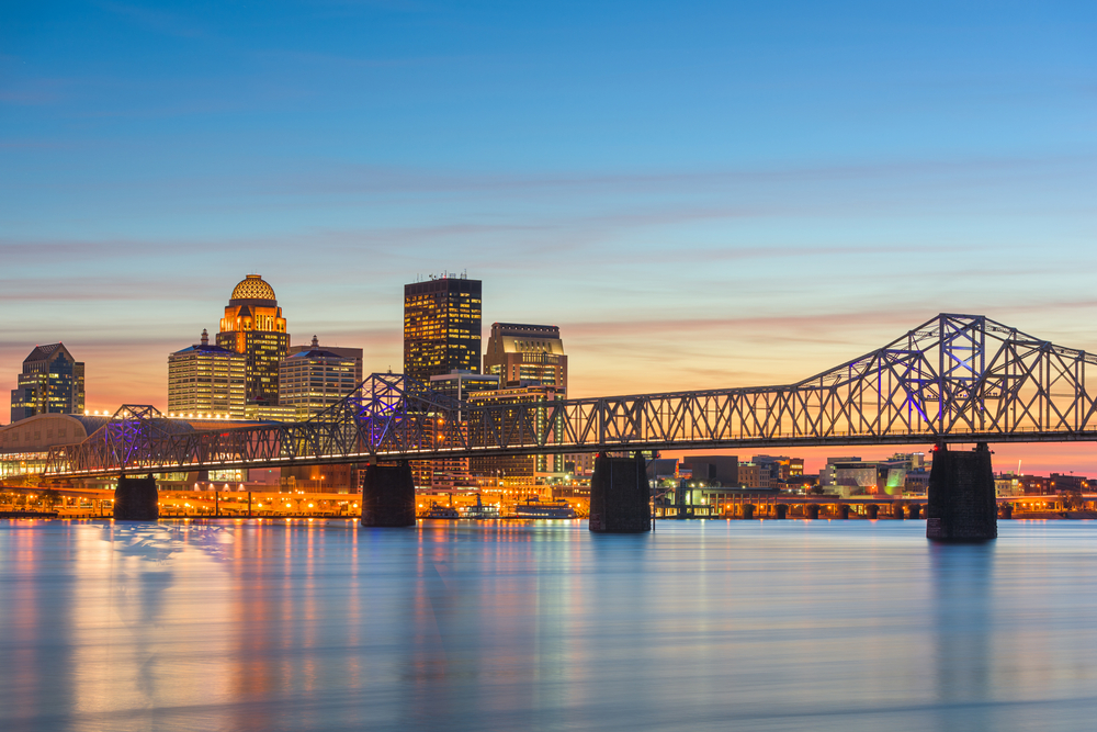 The skyline of downtown Louisville with several tall buildings stands against a blue sky with a bridge in the foreground.