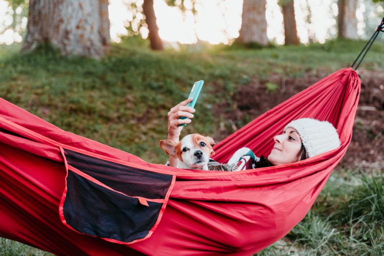 Woman lays in a hammock with her dog, looks at her phone
