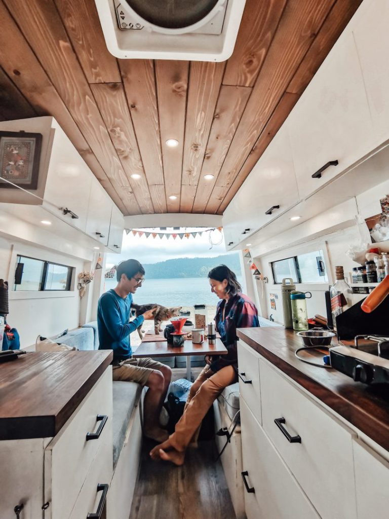 Couple sits at the table of their campervan with back doors open, a cat plays between them