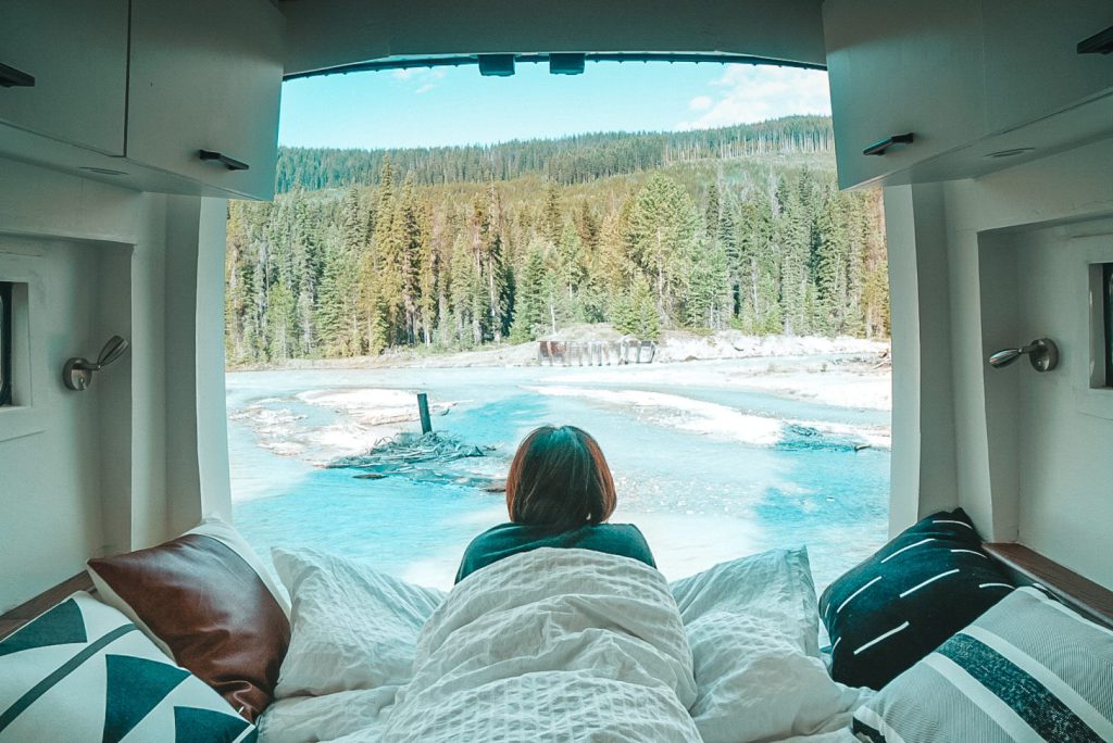 Woman lays in bed of her campervan with back doors open, looking out on a frozen lake