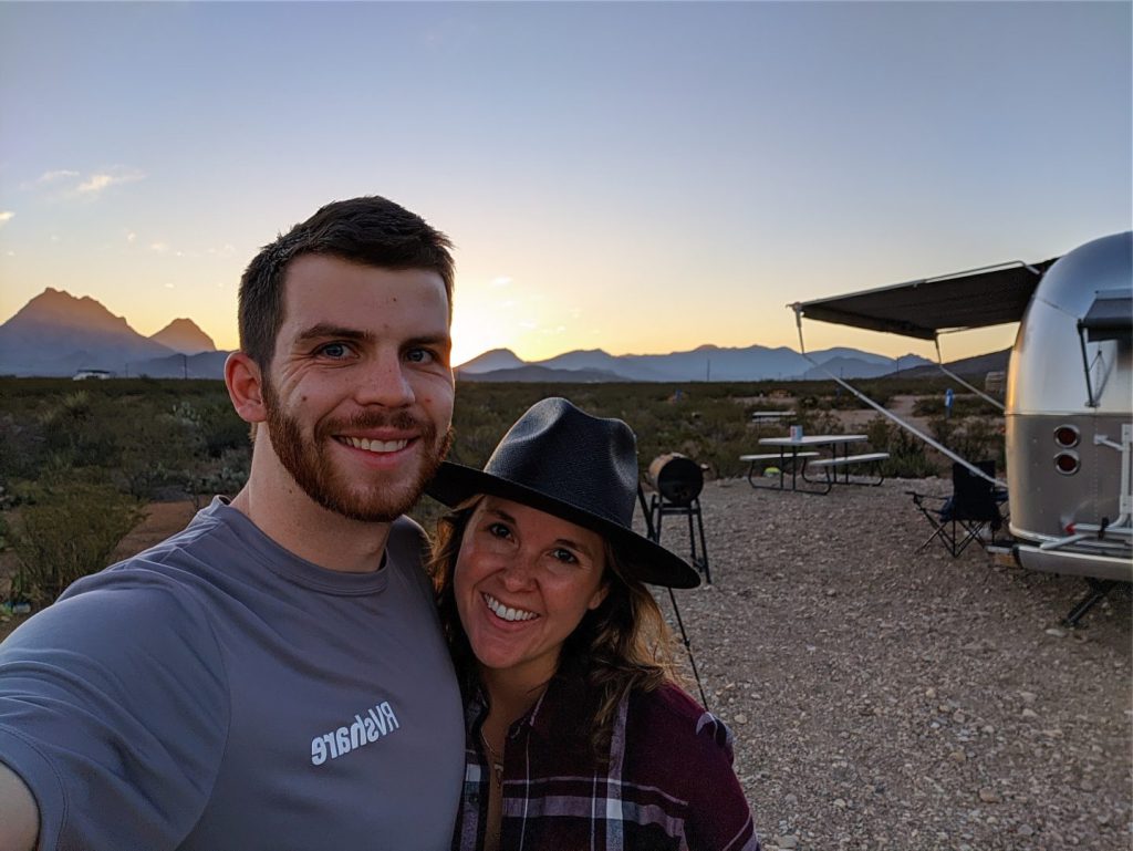 couple standing in front of an airstream trailer at sunset