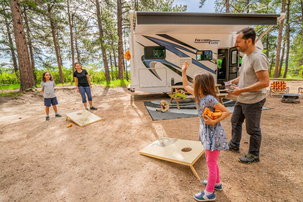 Mom, dad, and two children play corn hole/bag toss outside their parked Class C RV.
