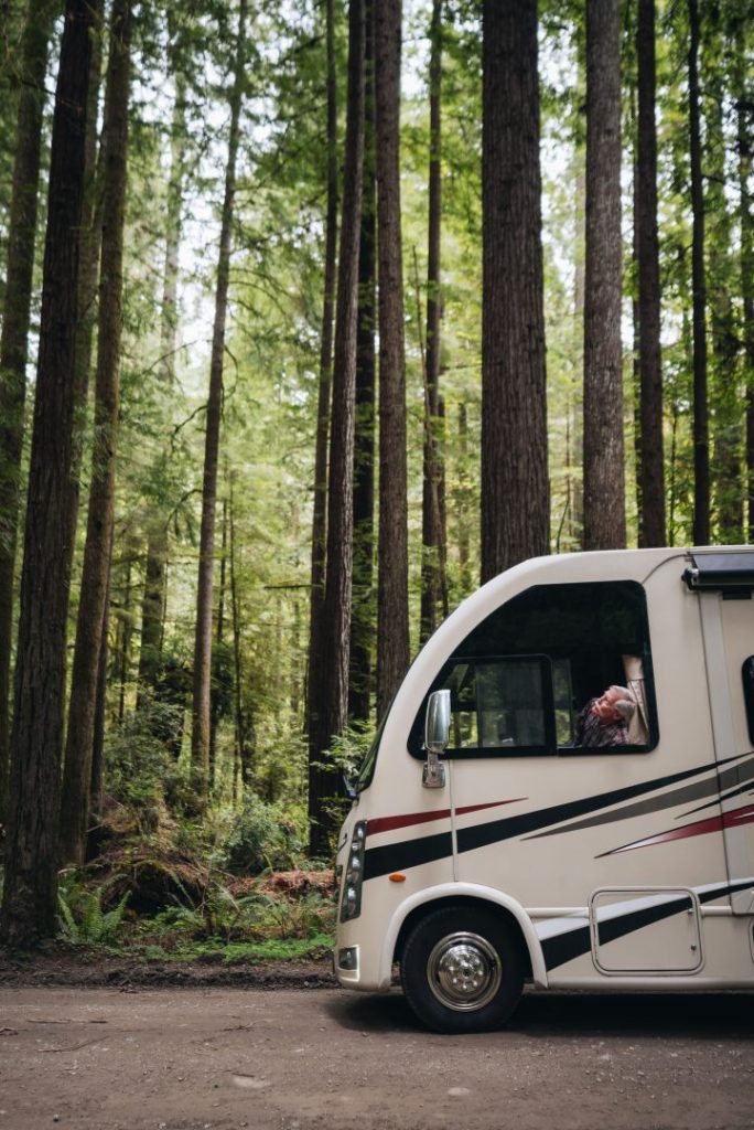 man looks out the window of a parked class a rv in the middle of the woods