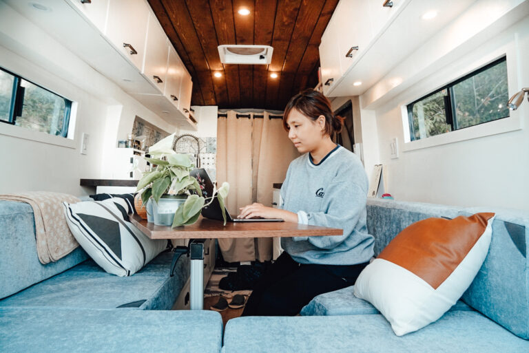 Woman sits at the table of her campervan working on her laptop