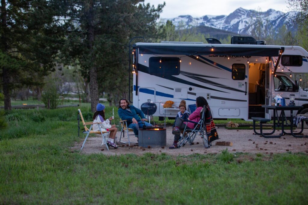 family sitting around a campfire at dusk with an RV in the background