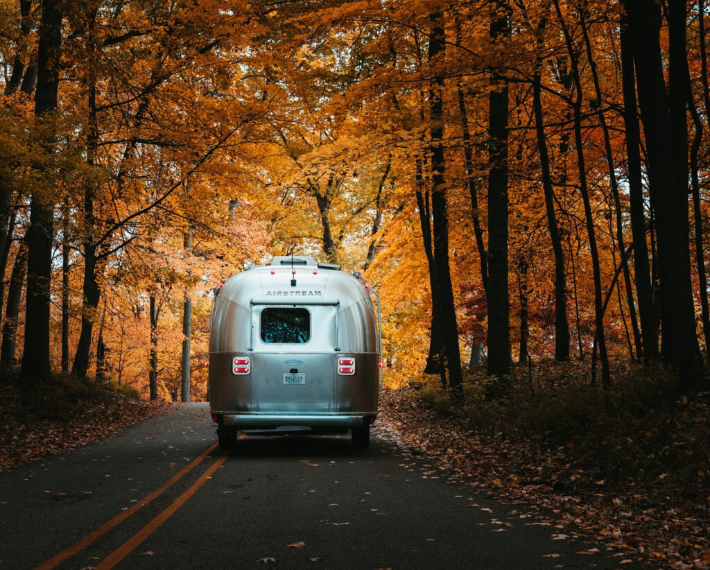 An Airstream trailer driving down a road lined with fall foliage