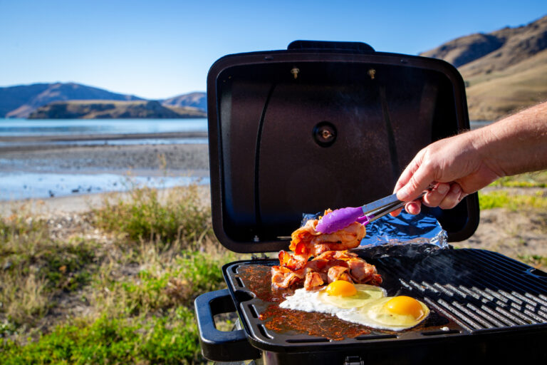 A person cooking breakfast on a portable grill with a lake in the background