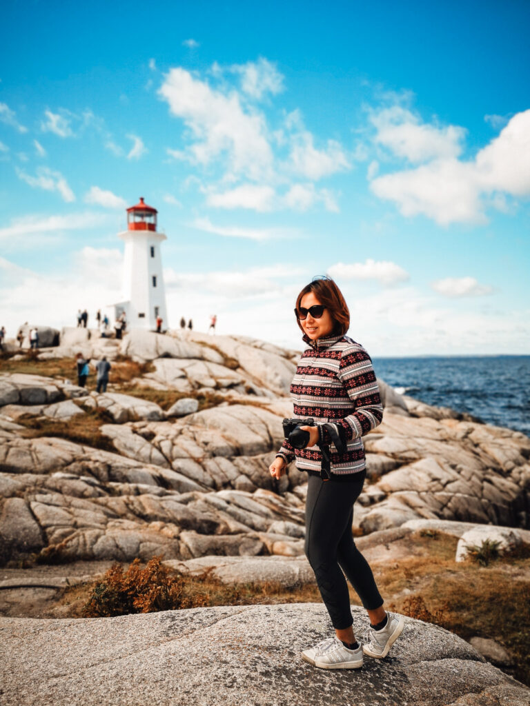 Woman stands on a rock by the sea holding a camera, lighthouse in the distance